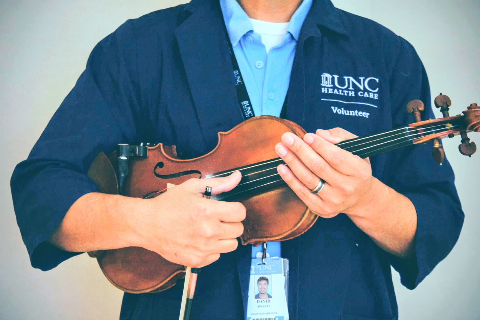 David holding his violin in his UNC Health Care Volunteer jacket with ID badge